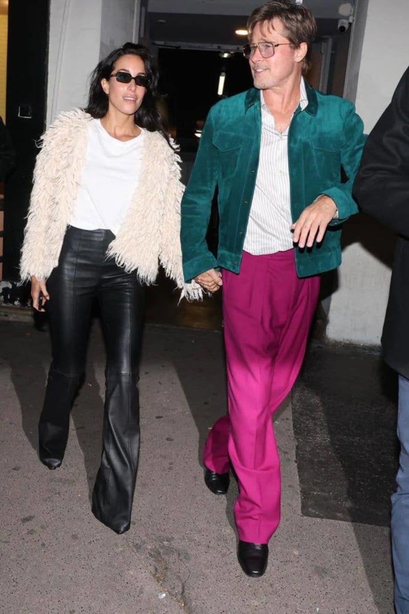 Two people walk together holding hands in an indoor corridor; woman wears sunglasses, cream jacket, and black pants; man wears teal jacket and magenta pants.