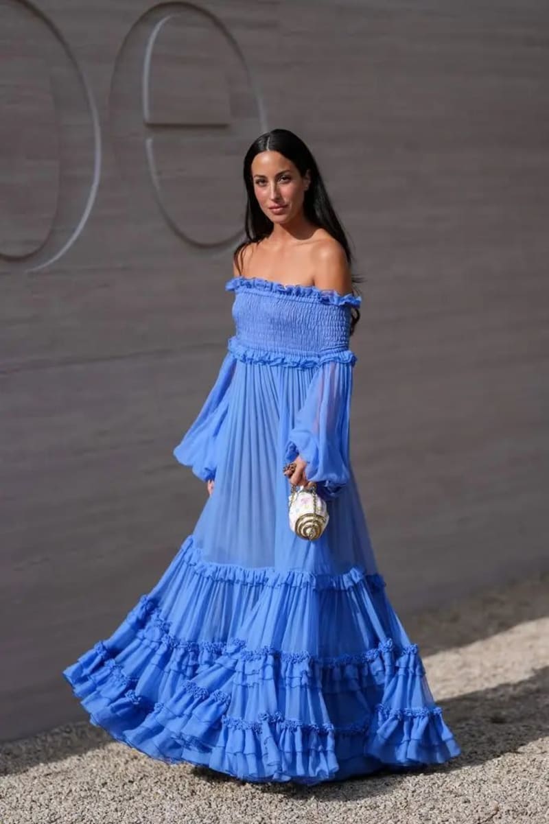 Woman in blue off-shoulder tiered maxi dress holding small ornate bag, outdoor event backdrop.