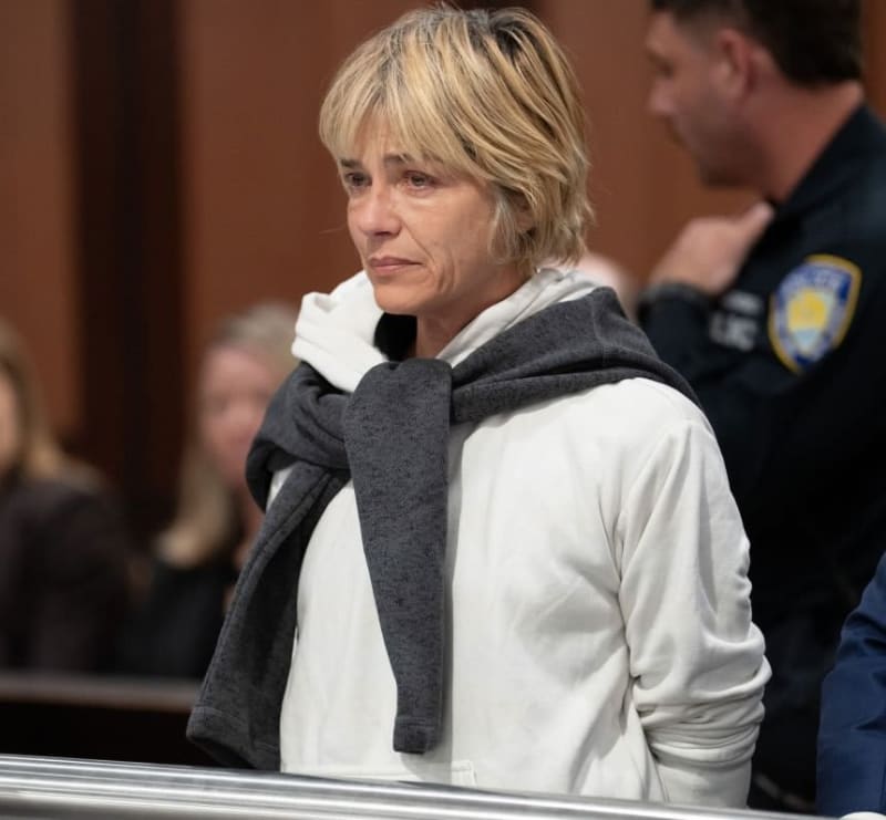 Woman in white hoodie and grey scarf standing in courtroom, officer visible behind.