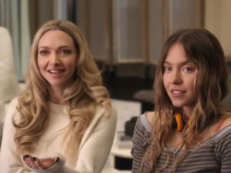Two women sitting indoors, one wearing a cream-colored sweater and the other in a striped gray shirt, both smiling at the camera.
