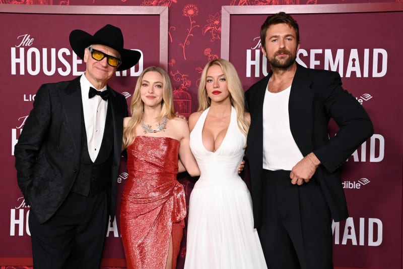 Man in black cowboy hat and tuxedo, woman in red gown, woman in white halter dress, man in black suit, at a red carpet premiere.