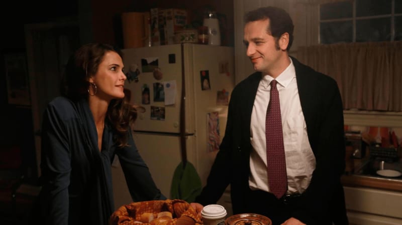 Two people in business attire having a conversation in a kitchen near a refrigerator with photos.