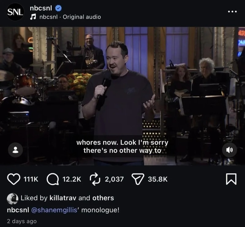 Man in dark t-shirt holding microphone on SNL stage, band visible behind him.