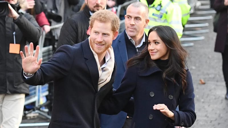 A man with reddish hair in a dark blazer waves to crowds while walking alongside a woman with long dark hair in a navy coat.