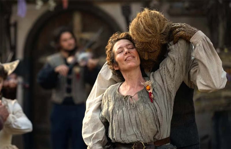 Woman in grey linen costume embraced by straw-masked creature, medieval village setting.