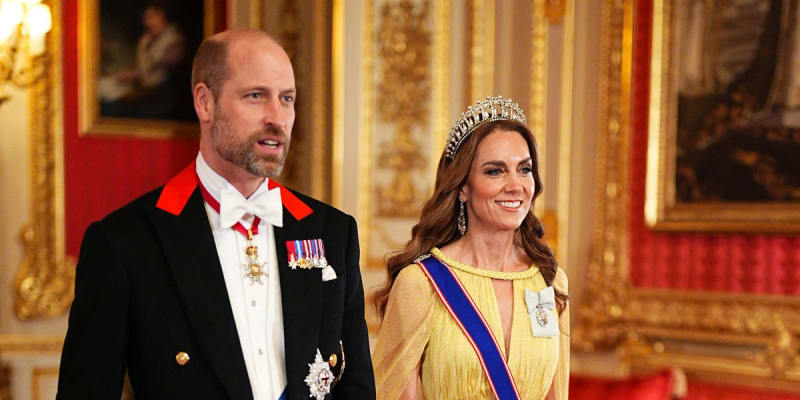 Two people in formal royal attire stand in an ornate palace room with gold-trimmed walls and red velvet furnishings.