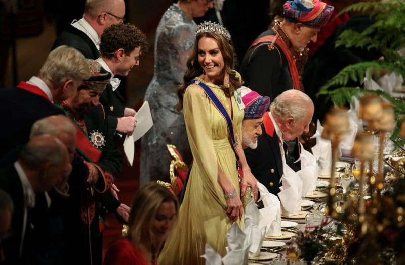 A woman in a cream gown and tiara stands at a formal state banquet surrounded by dignitaries in ceremonial dress and medals.