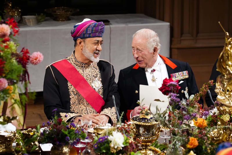 Two men in formal attire converse at an elegant state dinner, with ornate floral arrangements and gold candelabras adorning the table.