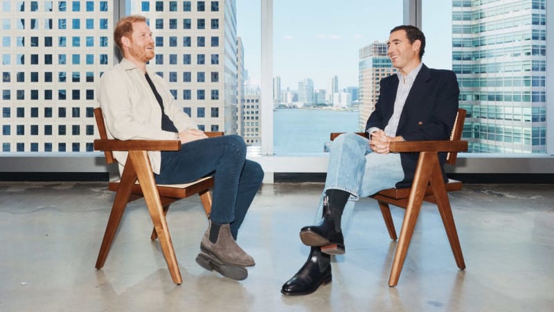 Two men sit in wooden chairs facing each other in a high-rise office with a city skyline and waterfront view behind them.