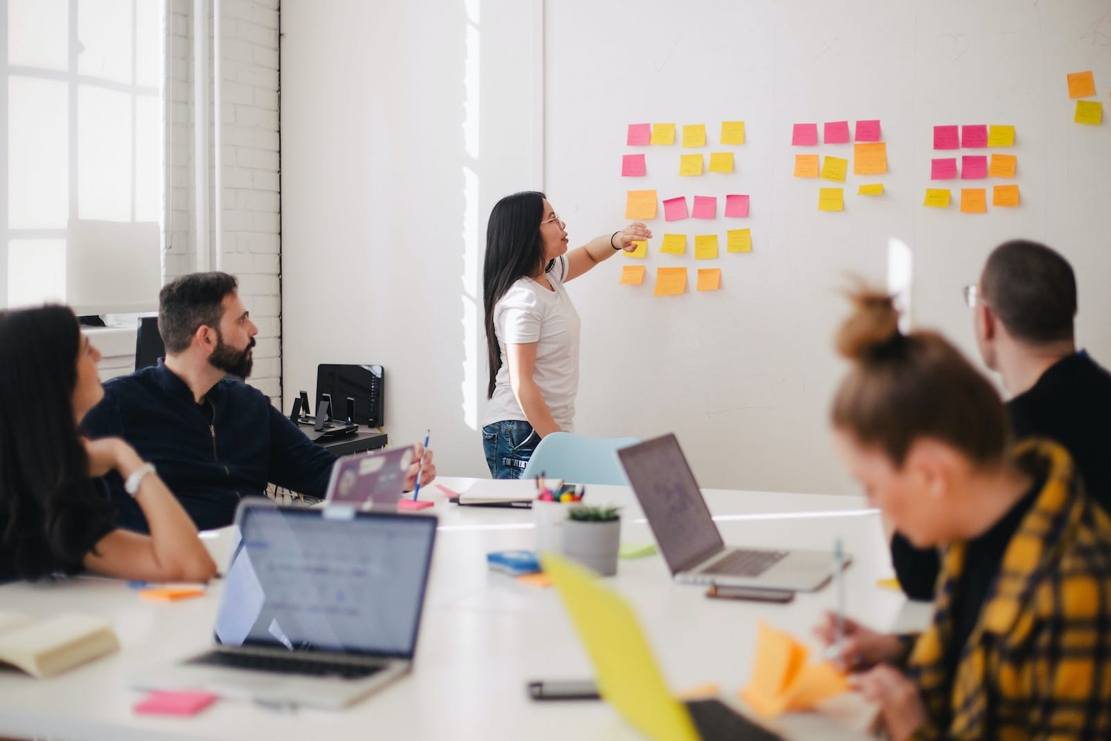 A team of four people around a desk in a meeting with one person talking through sticky notes that are stuck to a whiteboard