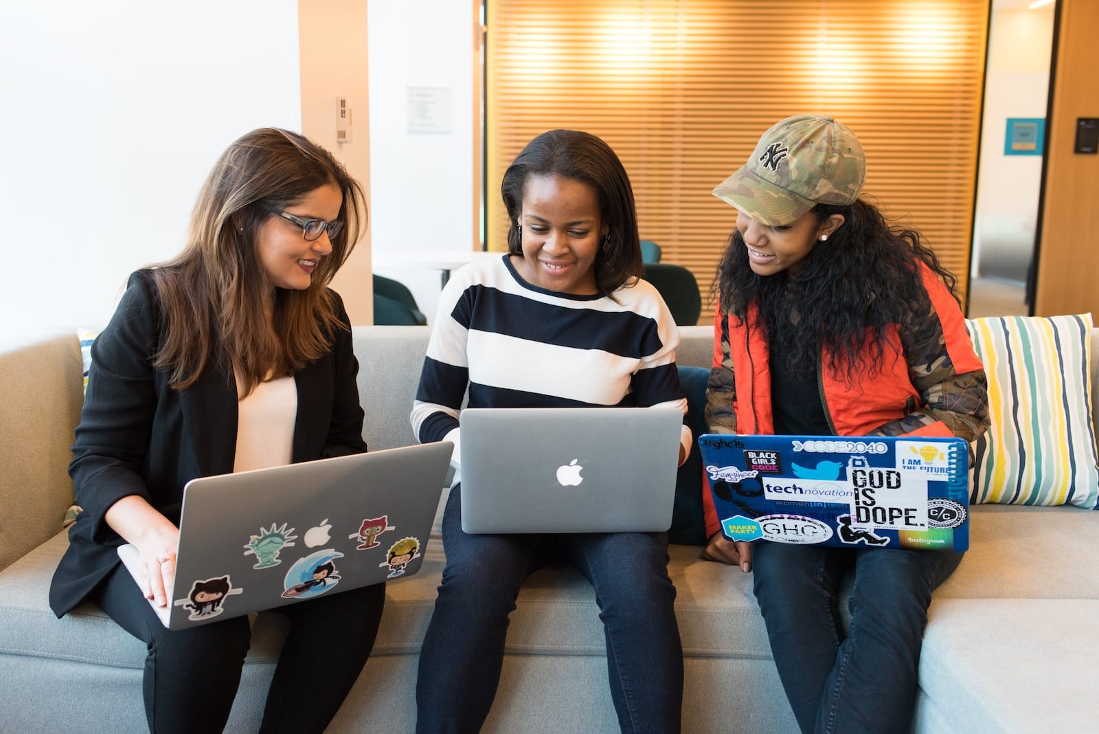 Three female co-workers sat on a sofa looking at the middle person's laptop