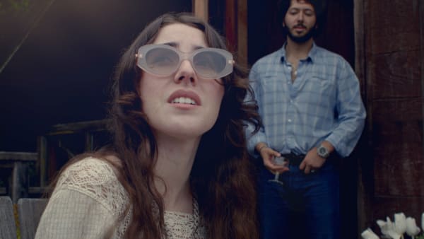 Production still: a woman in pale sunglasses in the foreground, a man watching from a doorway behind her.