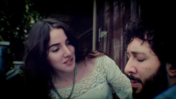Production still: two figures close together against weathered wood, faces turned toward each other.