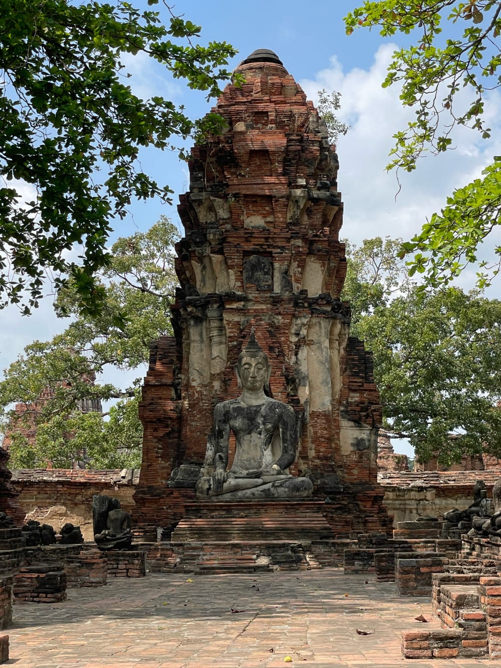 Bouddha assis devant un prang en brique encadré par des arbres dans le parc historique d'Ayutthaya