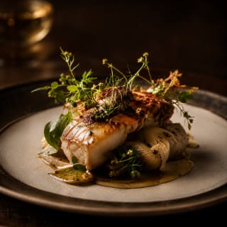 Filet de poisson blanc épais, doré sur le dessus, posé sur des quartiers de fenouil rôti et un jus clair, parsemé d'herbes fraîches et de petites fleurs jaunes, dans une assiette en grès beige mouchetée sur fond sombre.