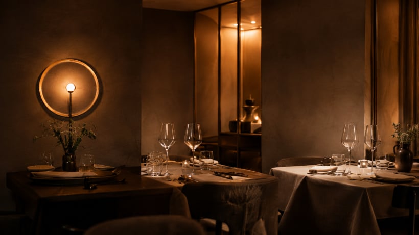 Salle du restaurant le soir, lumière tamisée : trois tables dressées de verres à vin, de couverts et de bougies, dont une nappée de lin clair, un bouquet champêtre dans un vase en grès et un grand luminaire circulaire allumé sur le mur d'enduit ocre.