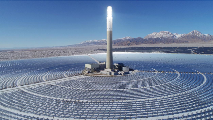 Aerial view of a concentrated solar thermal plant in arid country: thousands of mirrors arranged in concentric rings around a central receiver tower, with bare hills behind.