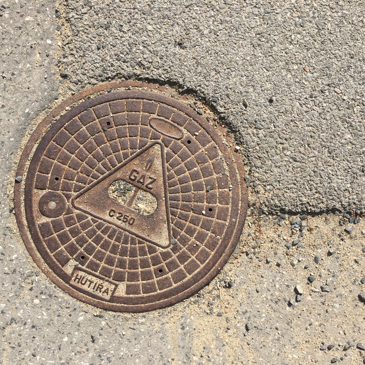 Kulatý litinový poklop, obklopený hrubým asfaltem a pískem, vykazuje výraznou geometrickou mřížku, která se radiálně roz in Praha, Czechia — CoverSpot