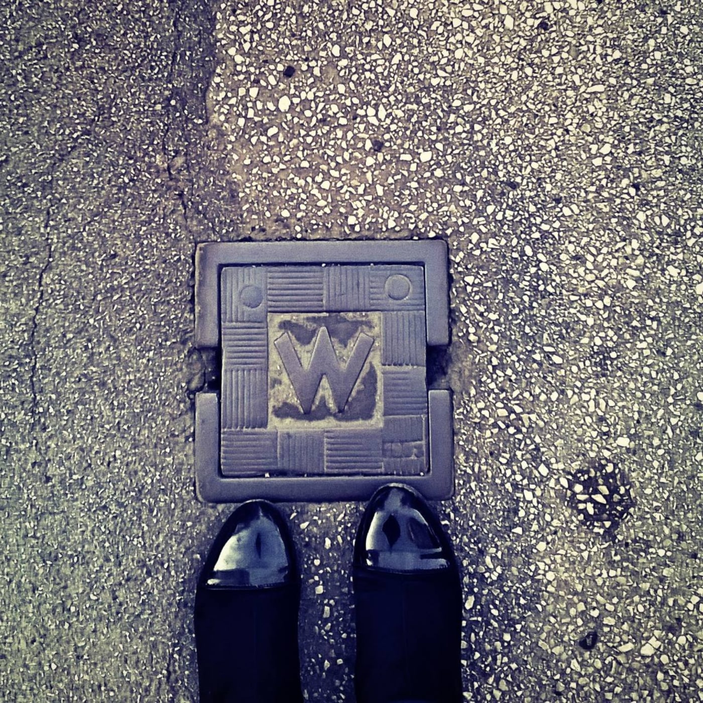 Na zrnitém šedém povrchu chodníku je zasazen čtvercový kovový prvek s výrazným písmenem 'M' uprostřed, obklopeným geomet in Vienna, Austria — CoverSpot
