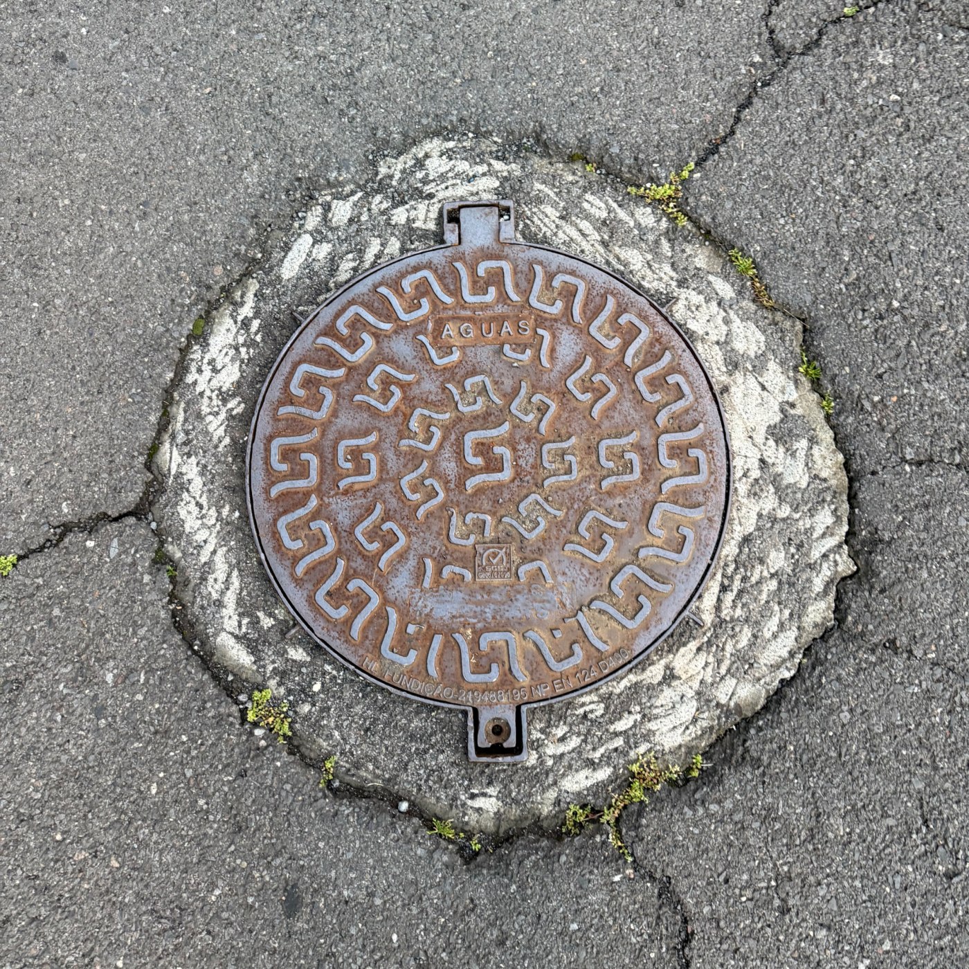 Opakující se geometrický vzor připomínající stylizované řecké klíče zdobí kruhový kovový povrch, s nápisem AGUAS uprostř i Curral das Freiras, Madeira, Portugal — CoverSpot
