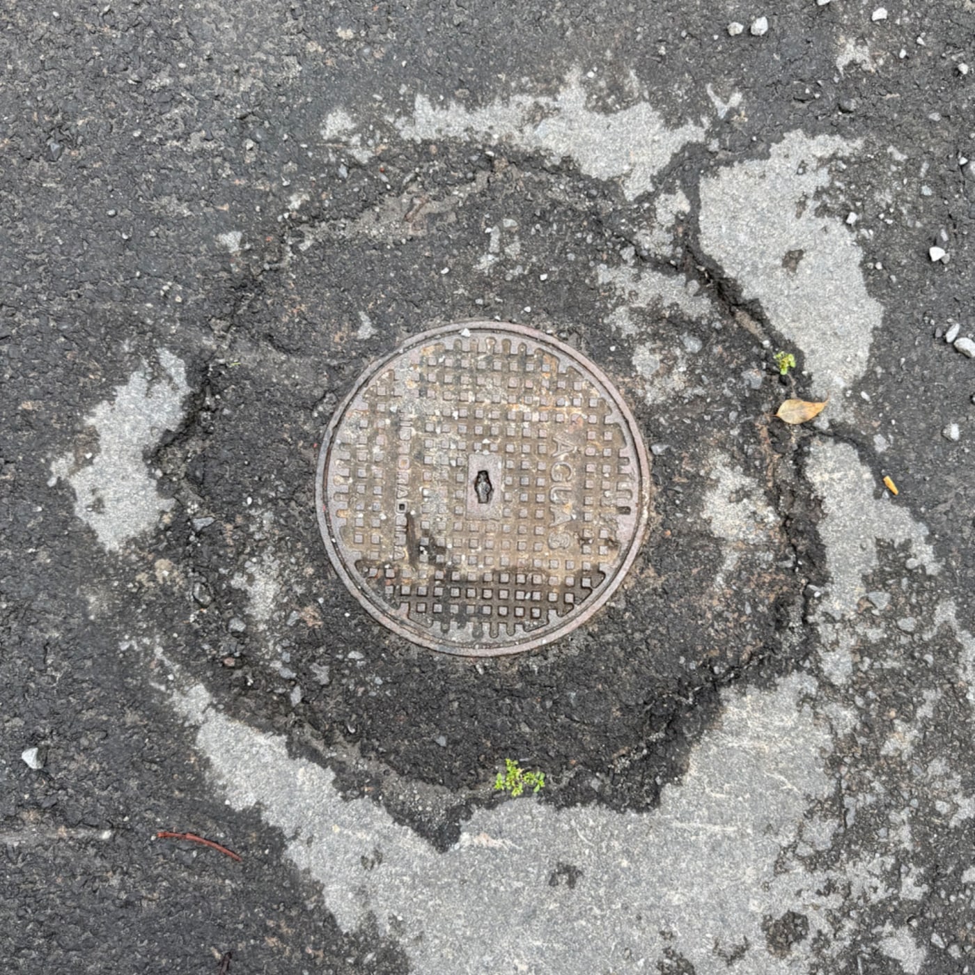 Kulatý poklop je vyroben z kovu a má na svém povrchu geometrický mřížkový vzor. Je zasazen do silně poškozeného asfaltu, in Santa Luzia, Madeira, Portugal — CoverSpot