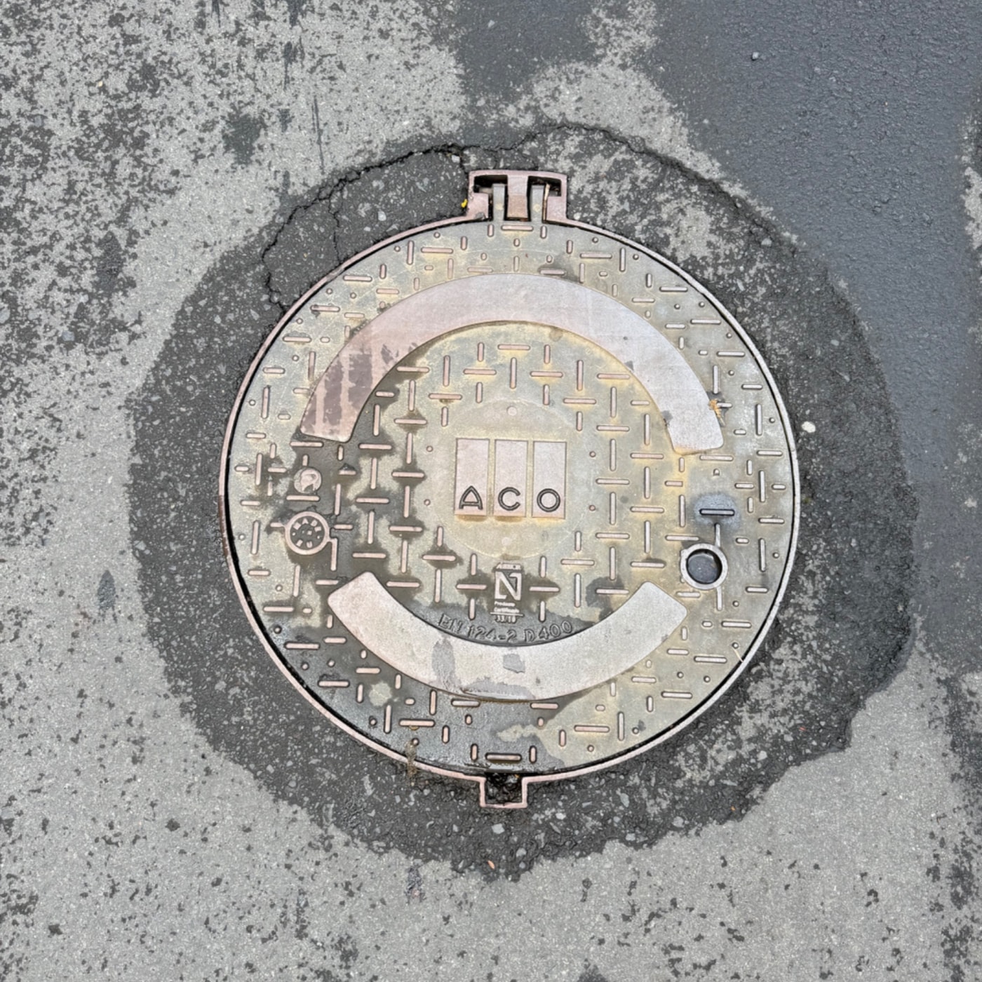 Jedná se o kulatý kovový poklop s texturovaným povrchem. Uprostřed je zřetelně viditelné logo "ACO" a geometrický vzor.  in Santa Luzia, Madeira, Portugal — CoverSpot