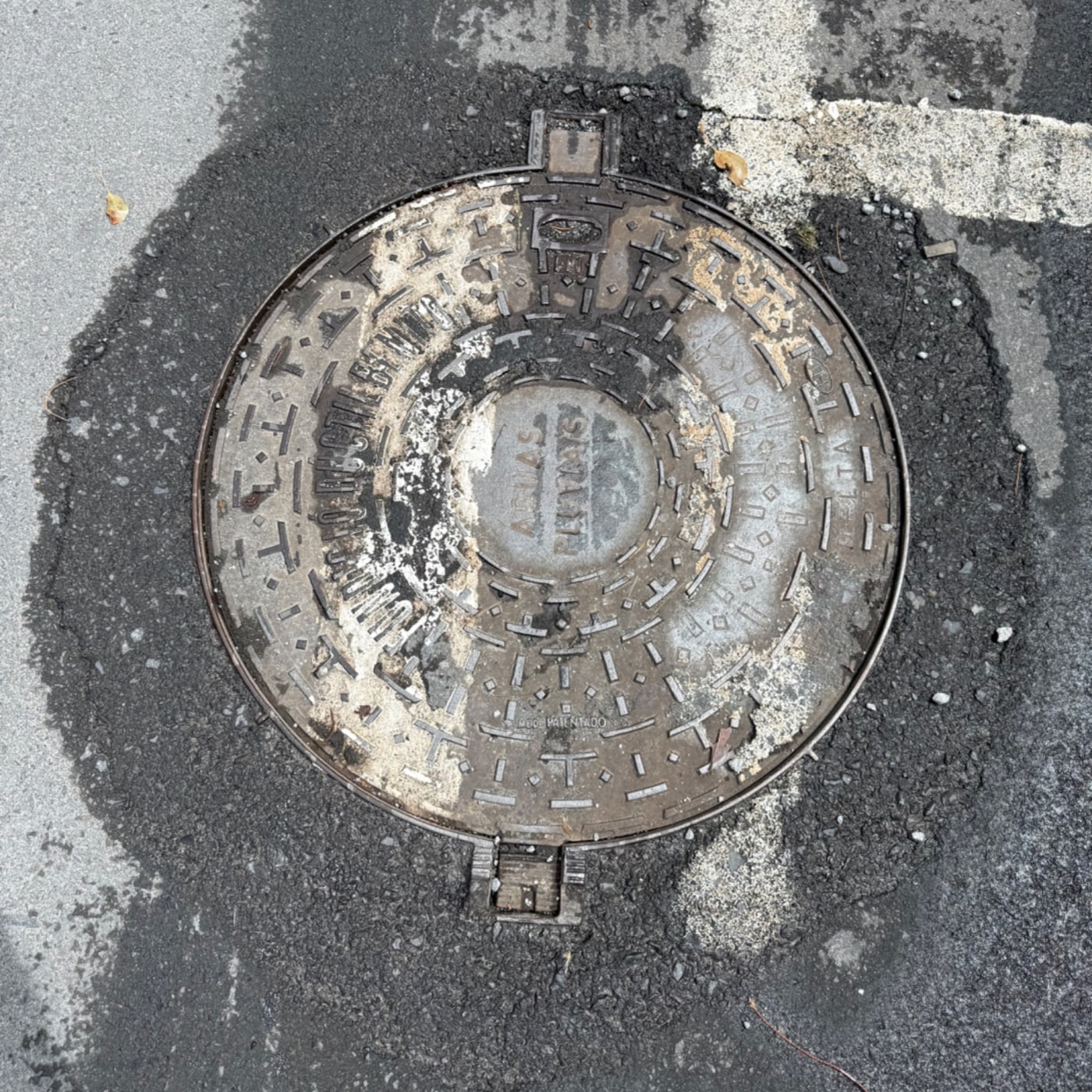 Kulatý poklop je vyroben z kovu a vykazuje známky opotřebení a znečištění. Je zasazen do asfaltu, kolem něj jsou viditel in Santa Luzia, Madeira, Portugal — CoverSpot