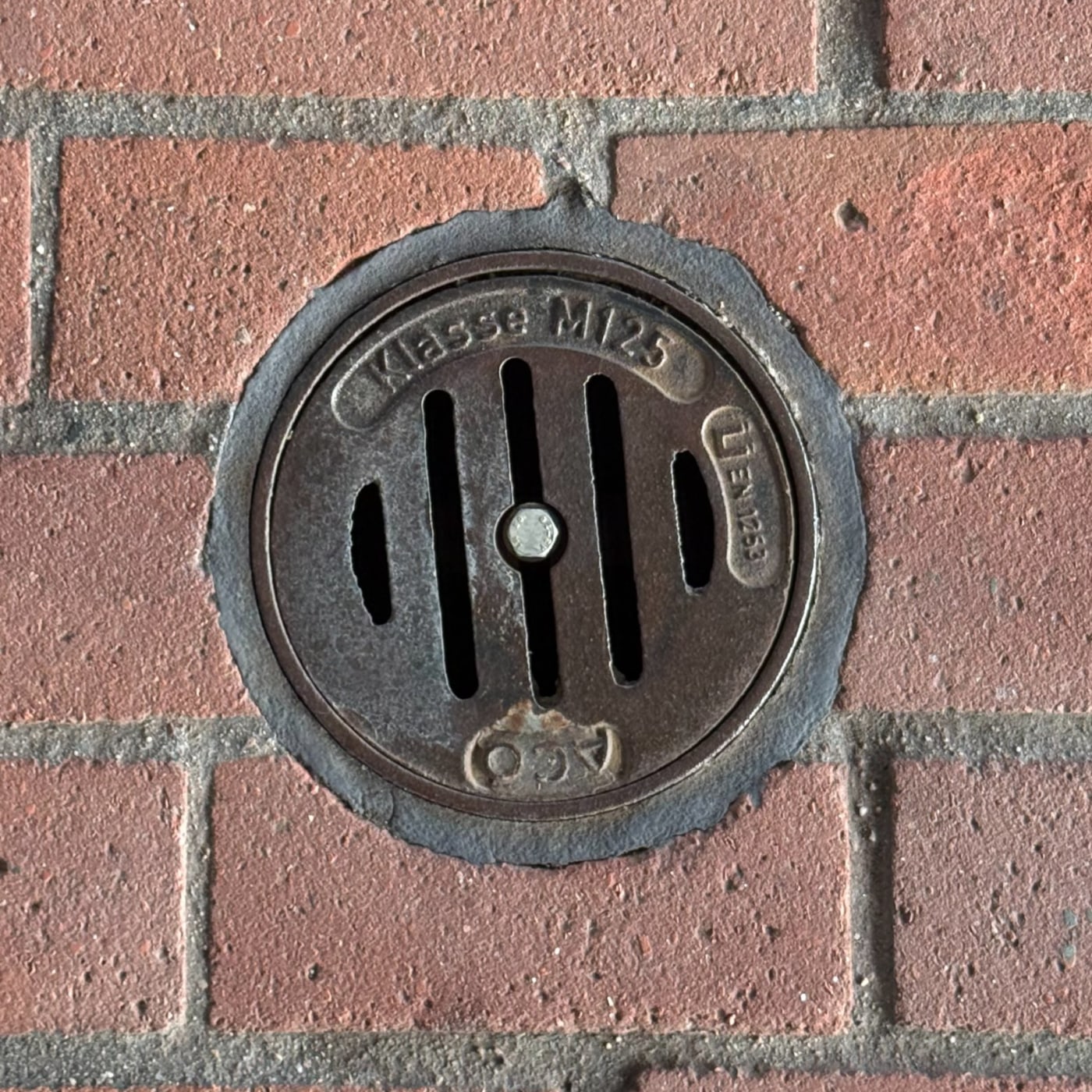 Na cihlové zdi je patrný kruhový odtokový prvek, jehož povrch nese zřetelné označení 'Klasse M125' a referenci na normu  in HafenCity, Free and Hanseatic City of Hamburg, Germany — CoverSpot