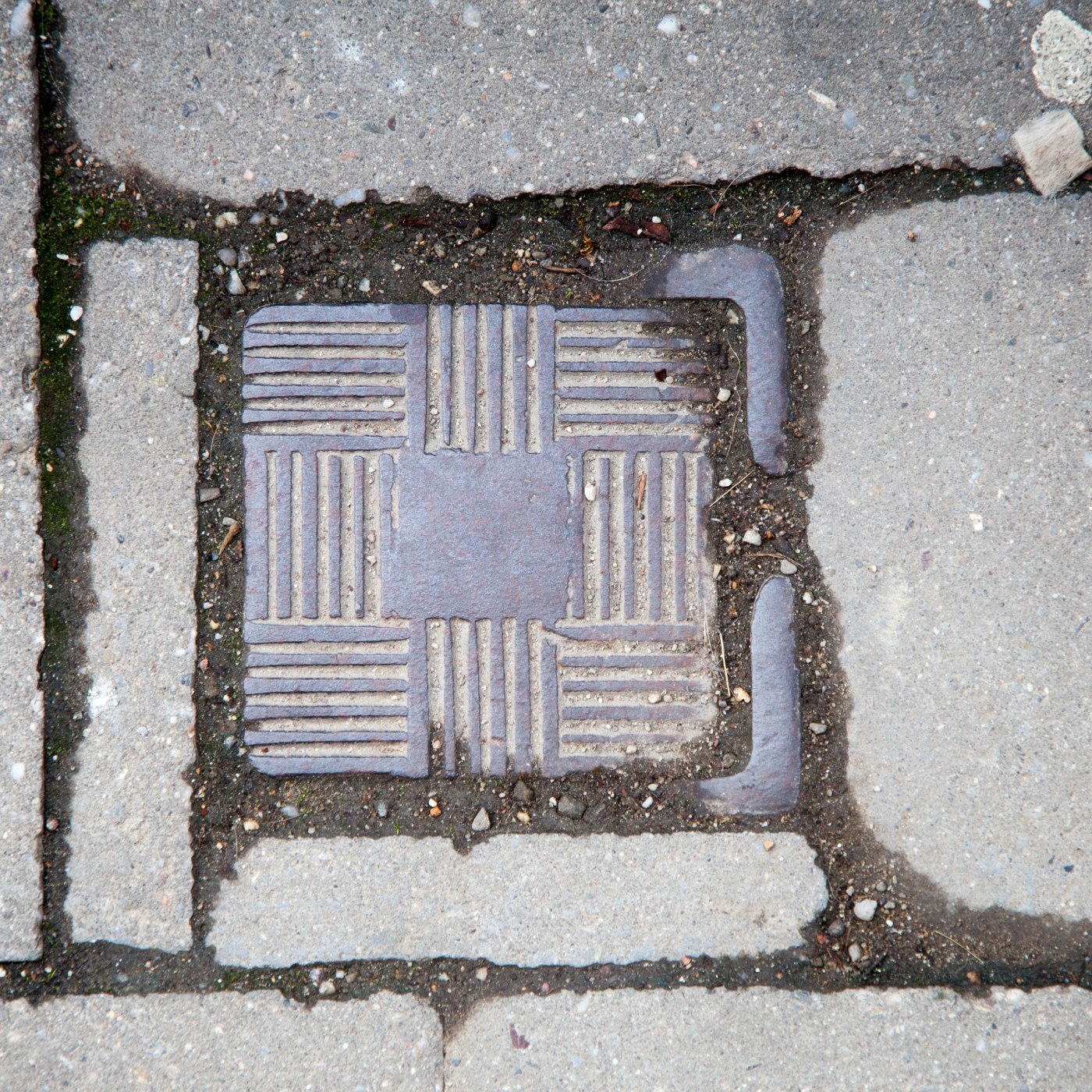 Čtvercová kovová deska, zasazená do betonové dlažby, vykazuje robustní geometrický vzor střídajících se horizontálních a in Prostějov, Czechia — CoverSpot