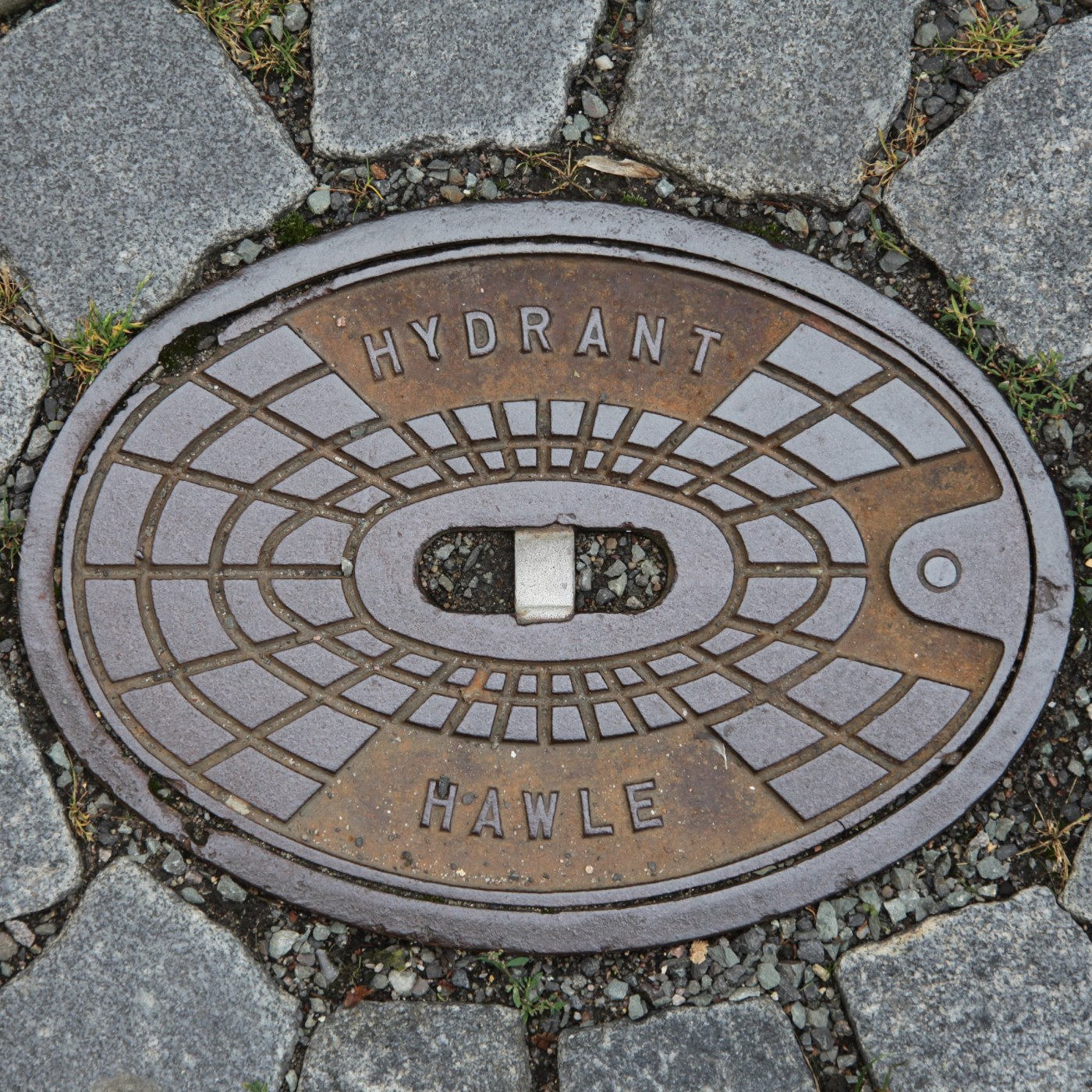 Výrazná patina rzi pokrývá kruhový litinový poklop, který je pečlivě zasazen do dlážděné cesty z hrubých kamenných koste in Hradec Králové, Czechia — CoverSpot