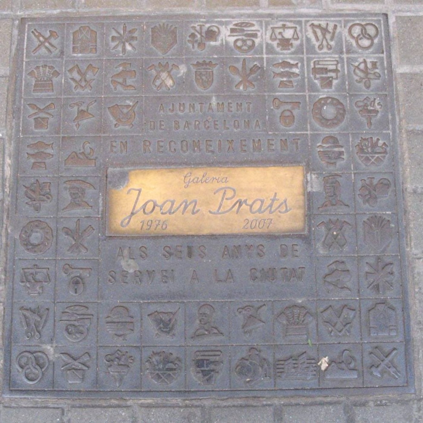 Na chodníku je zasazena čtvercová kovová deska, jejíž povrch je pokryt složitou mřížkou drobných, stylizovaných symbolů. in Barcelona, Catalonia, Spain — CoverSpot