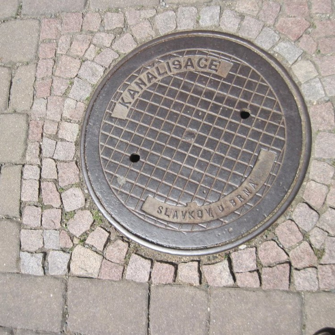Mřížkový vzor na povrchu tohoto exempláře litinové infrastruktury naznačuje jeho funkční účel. Nápisy 'KANALISACE' a 'SL — CoverSpot