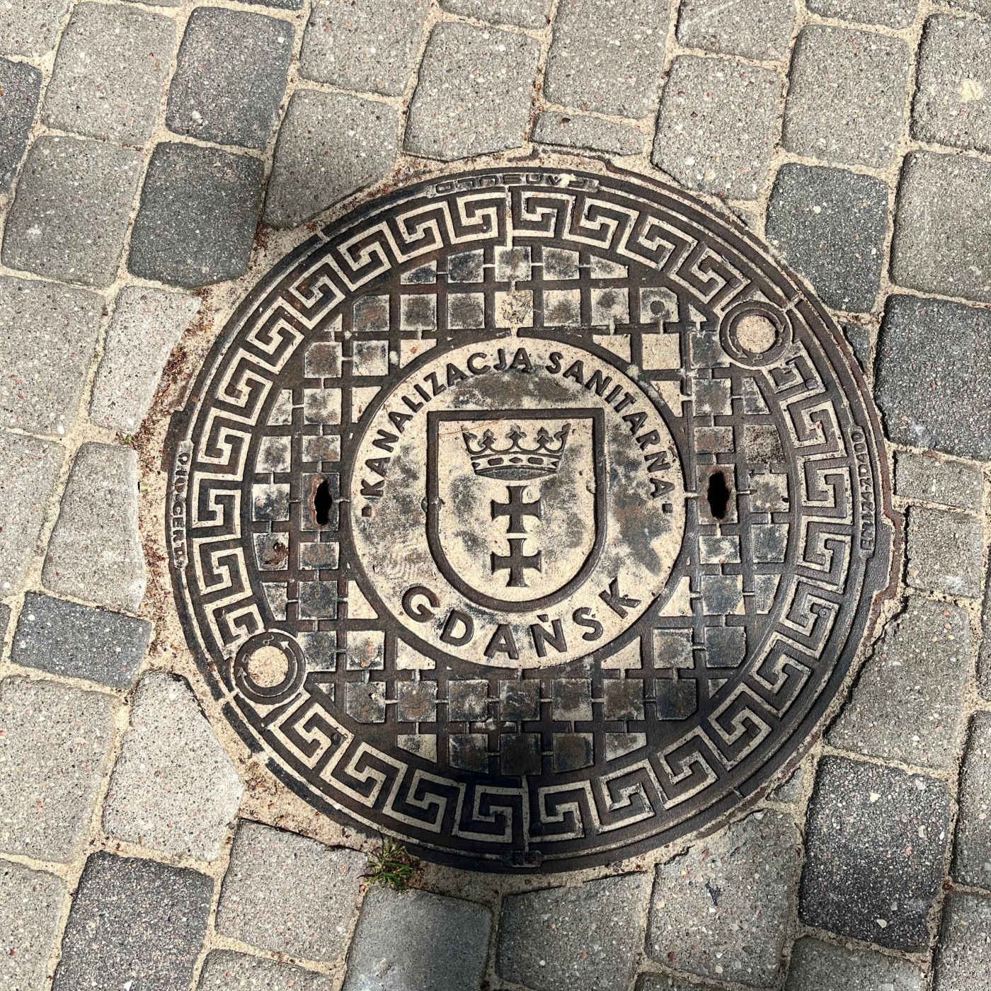 Kruhový kovový poklop je zasazen do dlážděné cesty z šedých a tmavších kostek. Jeho povrch zdobí složitý geometrický vzo in Gdańsk, Poland — CoverSpot