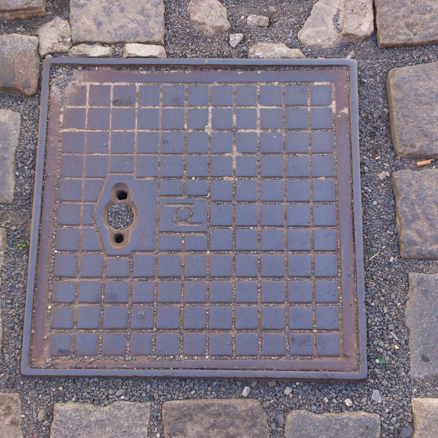 Mřížkovaný vzor pokrývá celou plochu čtvercového poklopu, který je obklopen drobným štěrkem a nepravidelnými dlažebními  in Olomouc, Olomoucký, Czechia — CoverSpot