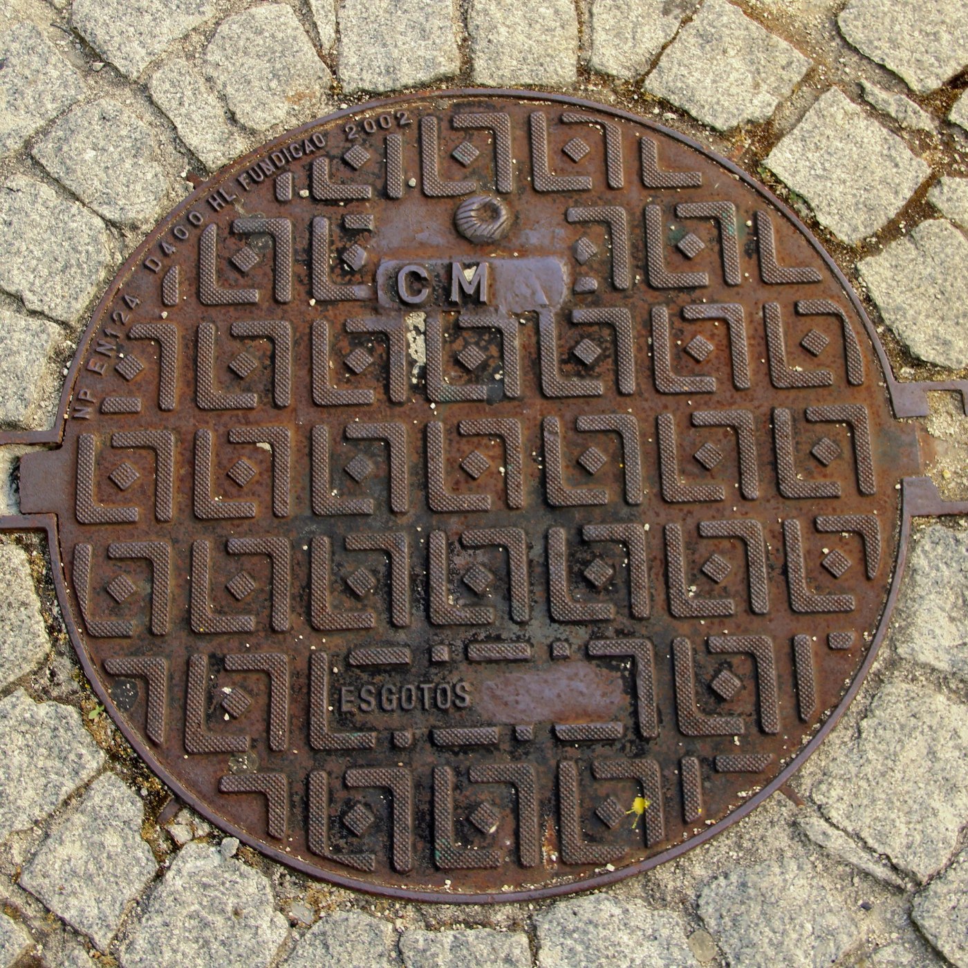 Výrazný geometrický vzor s opakujícími se prvky dominuje povrchu kruhového litinového poklopu, který je zasazen do dlážd in Lisbon, Portugal — CoverSpot