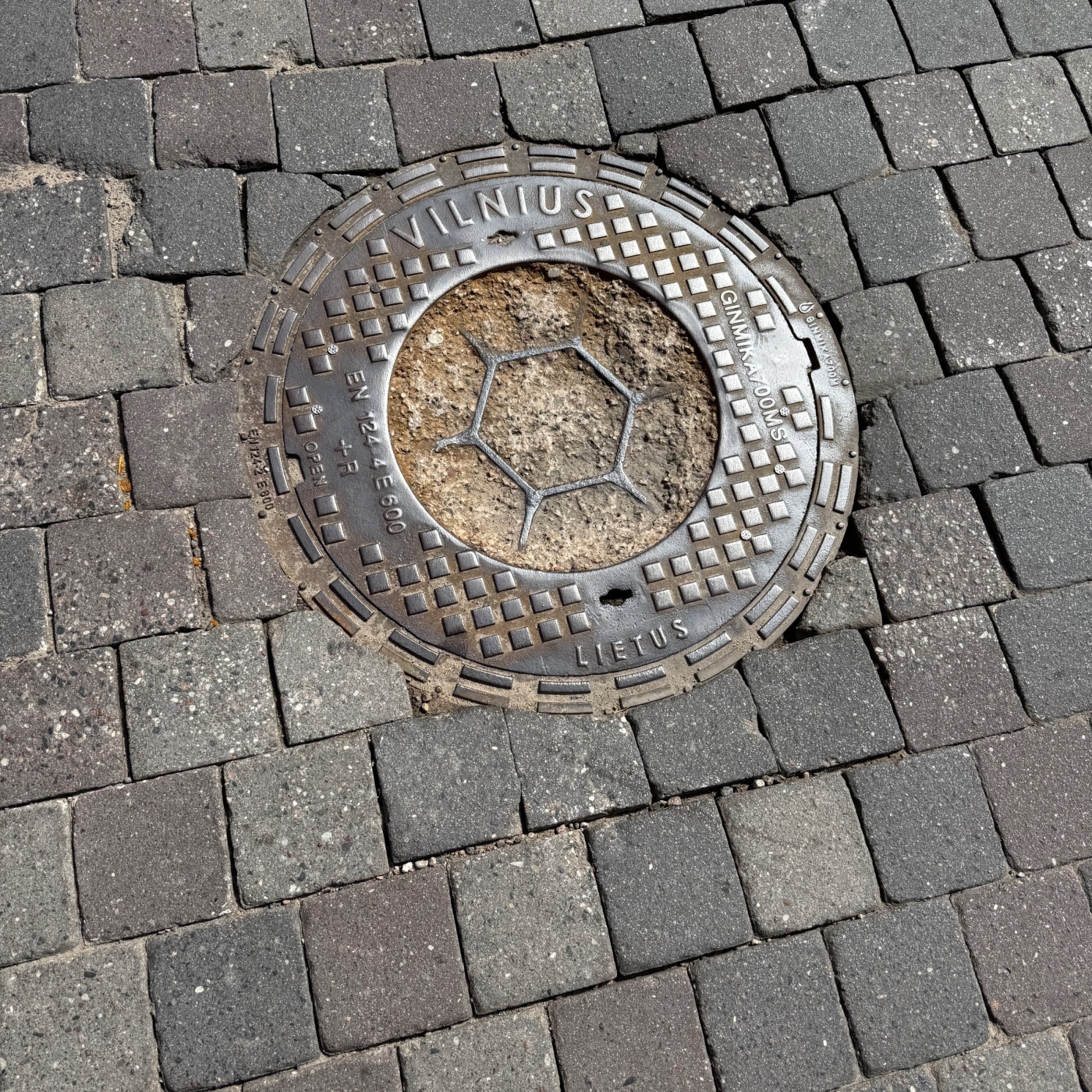 Kovový disk, zasazený do dlažby z tmavých kostek, nese nápis VILNIUS a LIETUS, což naznačuje jeho geografický původ a fu — CoverSpot