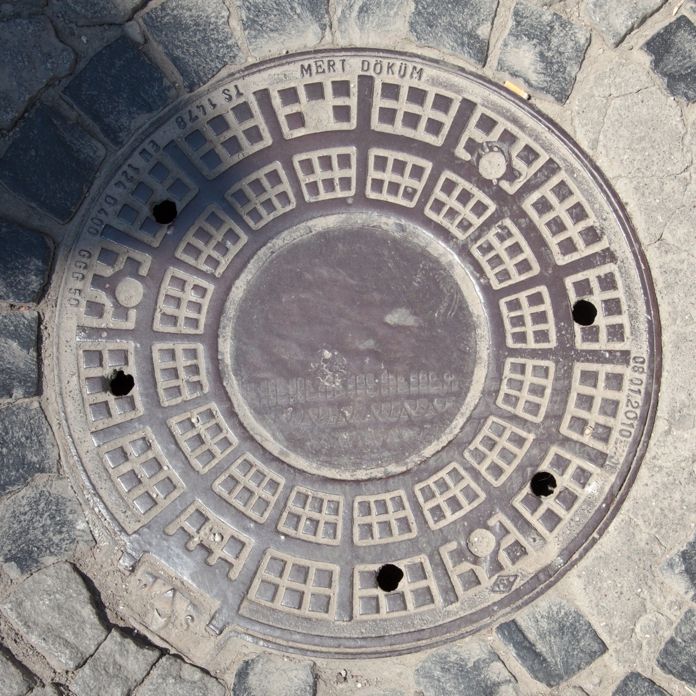 Dlážděná plocha z tmavých kostek a světlého betonu obklopuje kruhový poklop, jehož povrch zdobí opakující se geometrický in Istanbul, Turkey — CoverSpot