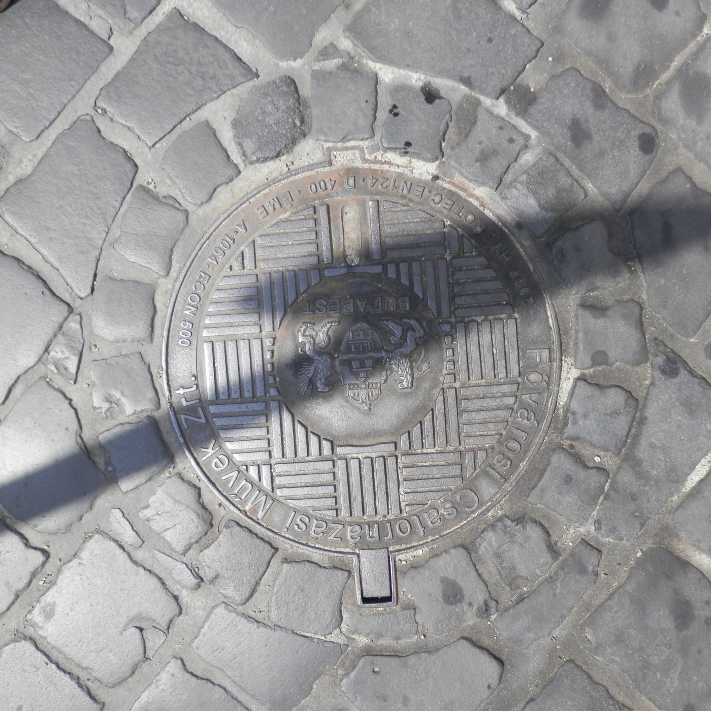 Dlažba z nepravidelných kamenných kostek obklopuje tento kruhový exemplář městské infrastruktury. Uprostřed je zřetelný  in Budapest, Hungary — CoverSpot