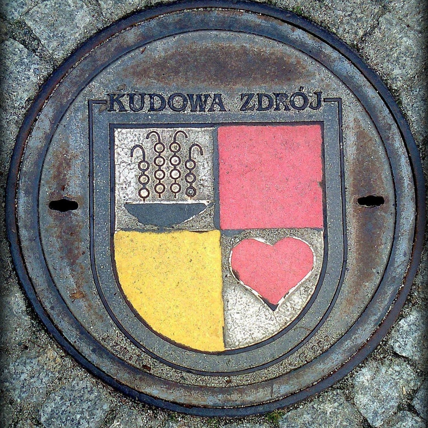 Centrální prvek tvoří heraldický štít města Kudowa Zdrój, pečlivě vlitý do kovového povrchu. V jeho čtyřech polích lze p — CoverSpot