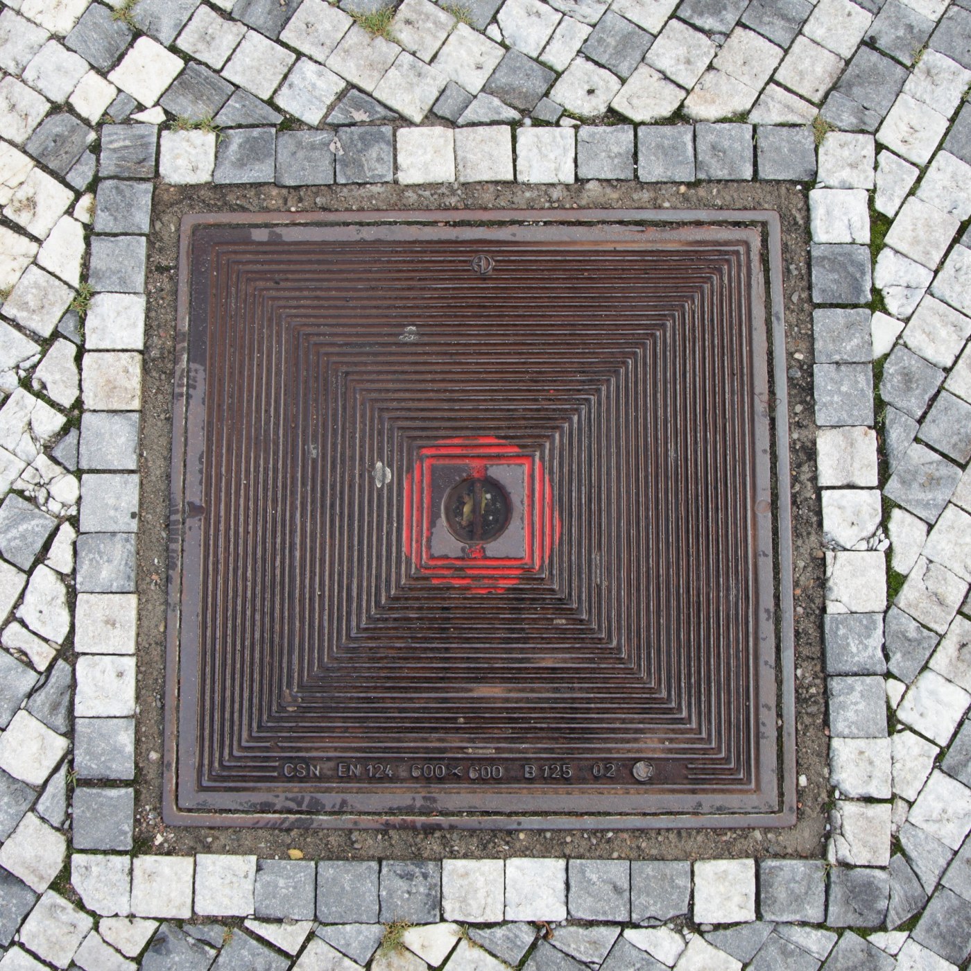 Koncentrické čtvercové drážky dominují tomuto exempláři městské infrastruktury, s výrazným červeným čtvercem uprostřed,  in Hradec Králové, Czechia — CoverSpot