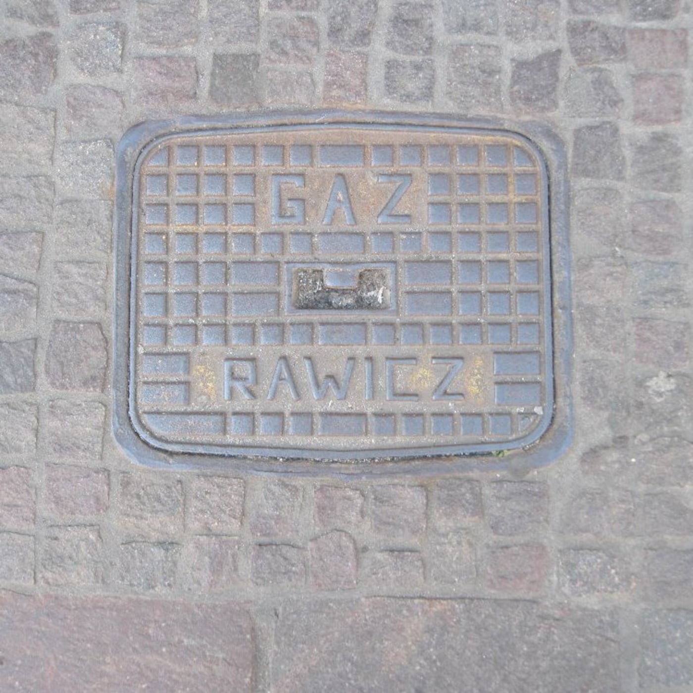 Mřížkovaný povrch tohoto exempláře z litiny nese zřetelné nápisy "GAZ" a "RAWICZ", což naznačuje jeho funkční zařazení.  — CoverSpot