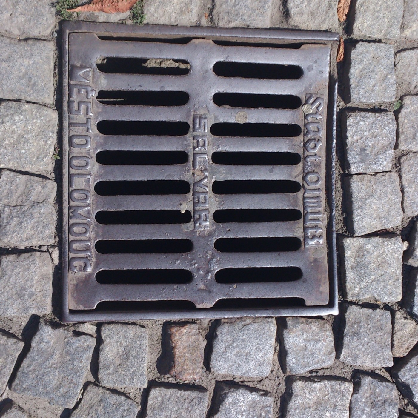 Hrubá textura litinového roštu, zasazeného do dlažebních kostek, odhaluje nápisy odkazující na město Olomouc a značku 'B in Olomouc, Czechia — CoverSpot