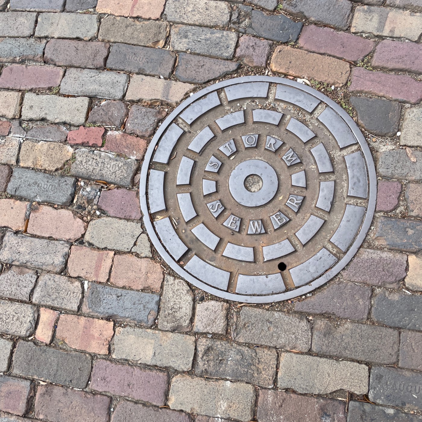 Kruhová kovová deska, zasazená do dlážděné cesty z cihel různých odstínů, nese výrazný nápis 'STORM SEWER'. Její povrch  in Manhattan, New York, United States — CoverSpot