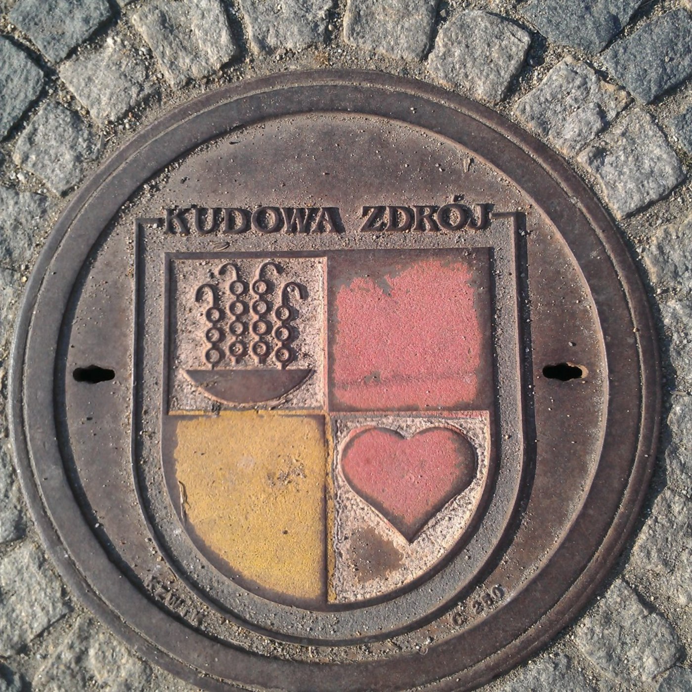 Zřetelný nápis 'KUDOWA ZDRÓJ' dominuje horní části tohoto kruhového exempláře, obklopeného dlažebními kostkami. Centráln in Kudowa-Zdrój, Poland — CoverSpot