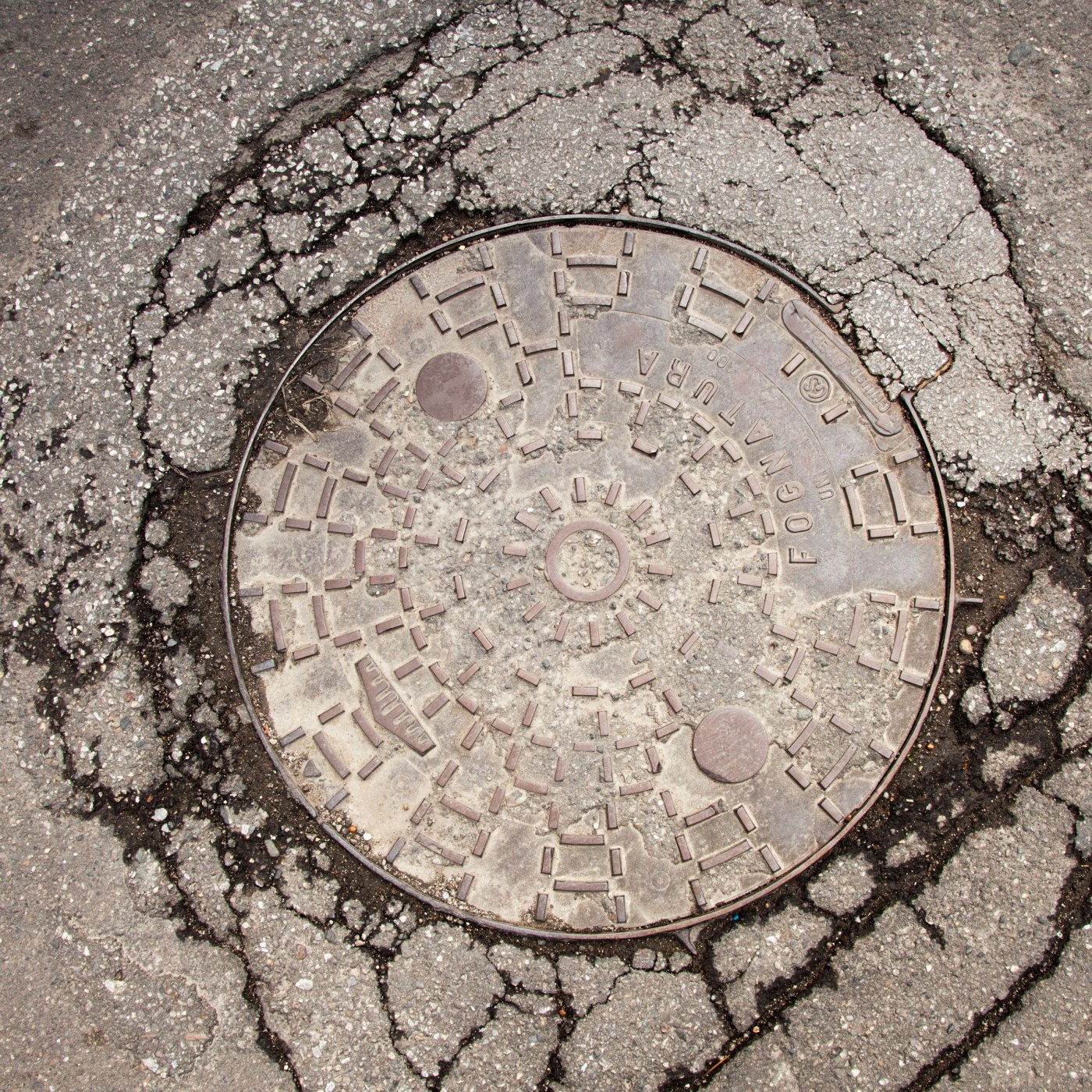 Kruhový poklop je zasazen do silně popraskaného asfaltu, což naznačuje značné opotřebení okolní vozovky. Jeho povrch zdo in Cefalù, Italy — CoverSpot