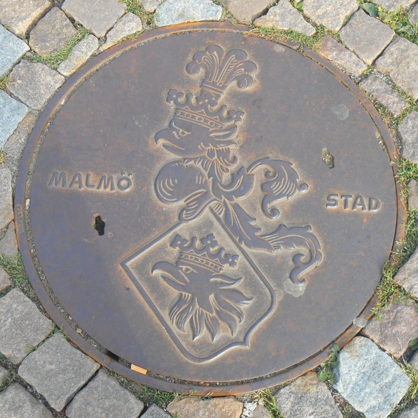 Tento kruhový poklop zdobí výrazný městský erb s korunovaným orlem, který se majestátně tyčí nad štítem s dalším orlem.  in Malmö, Sweden — CoverSpot