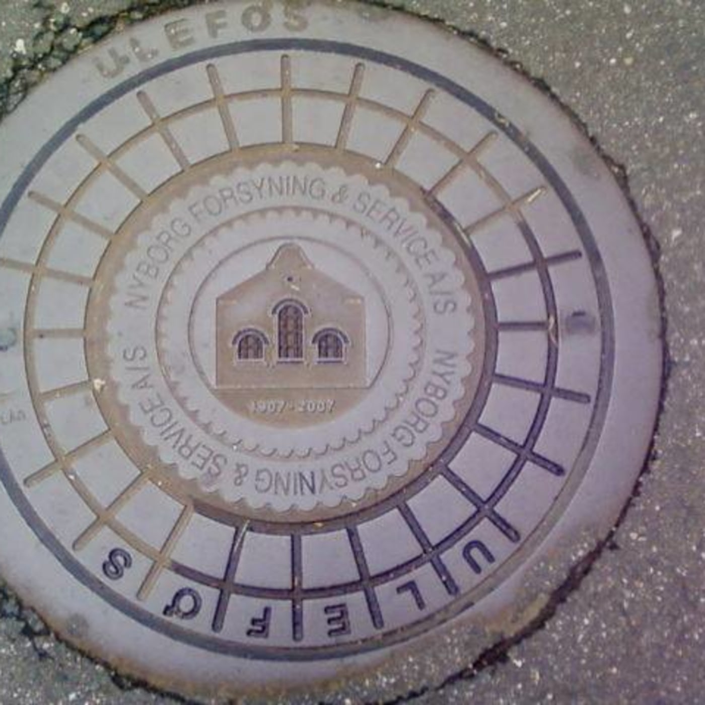 A round, grey cast iron manhole cover featuring a central emblem of a building, likely a historical utility structure. I in Poděbrady, Central Bohemia, Czechia — CoverSpot