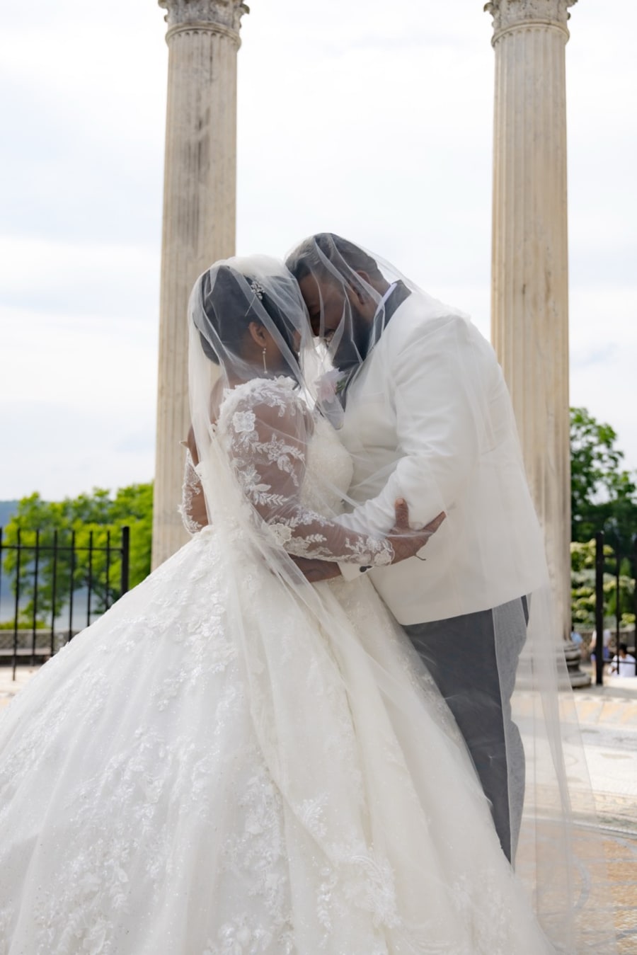Bride and groom kissing under a sheer veil between stone columns