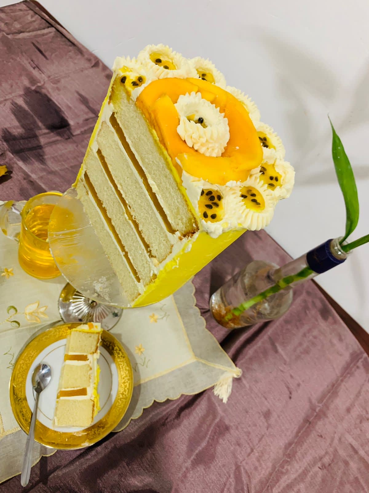 A multi-layered tropical sponge cake featuring clean geometric layers of passion fruit and mango gelee filling, frosted with smooth yellow buttercream and topped with fresh mango slices and passion fruit pulp on a tall glass stand.
