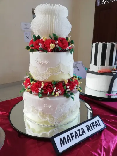 A classic three-tiered exhibition wedding cake featuring complex Lambeth method royal icing stringwork, delicate extension laces, dense red sugar rose borders, and an ornate white structural topper displayed on a mirror base.